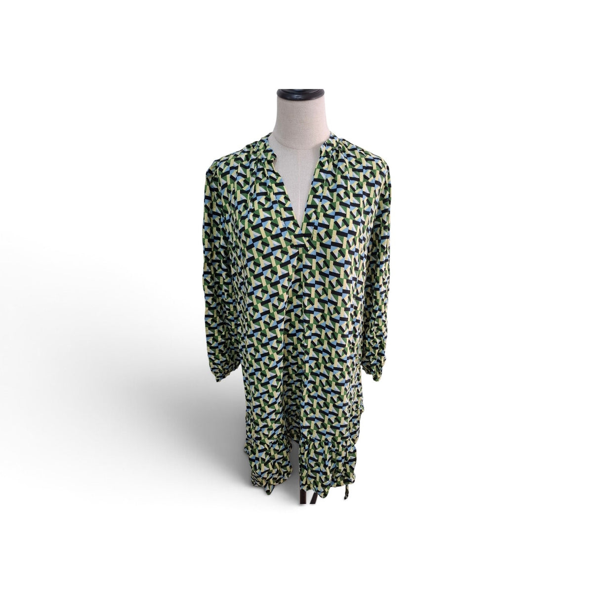 A long-sleeve V-neck tunic dress with an all-over abstract geometric pattern in green, blue, and yellow on a white base, displayed on a mannequin torso against a white background.