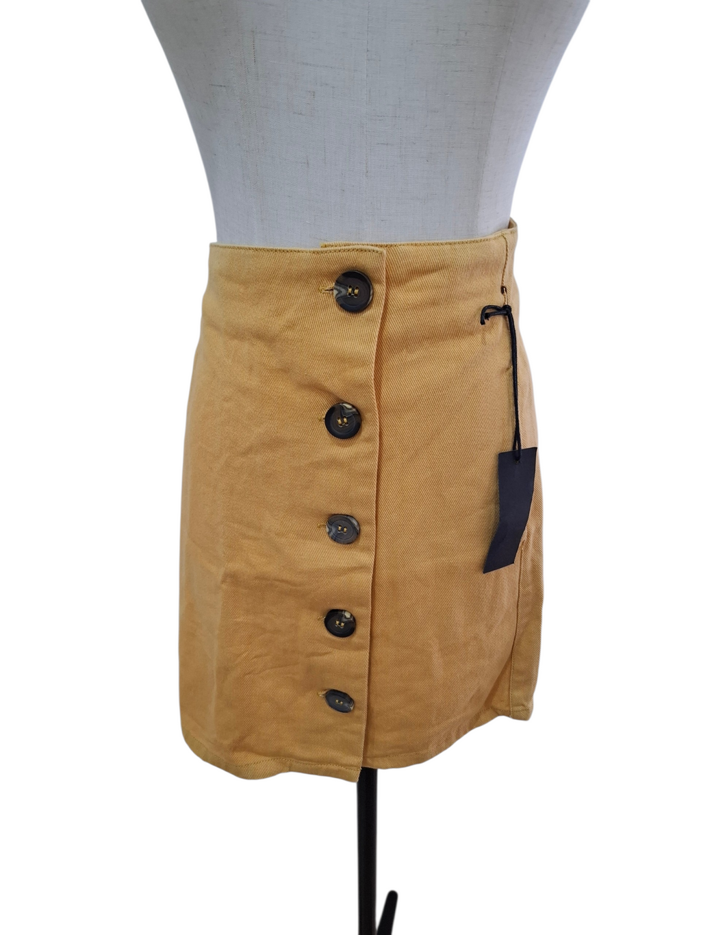 A mustard yellow mini skirt with a high waist, featuring a front button closure and a side pocket with a black tag, displayed on a white dress form mannequin against a white background.