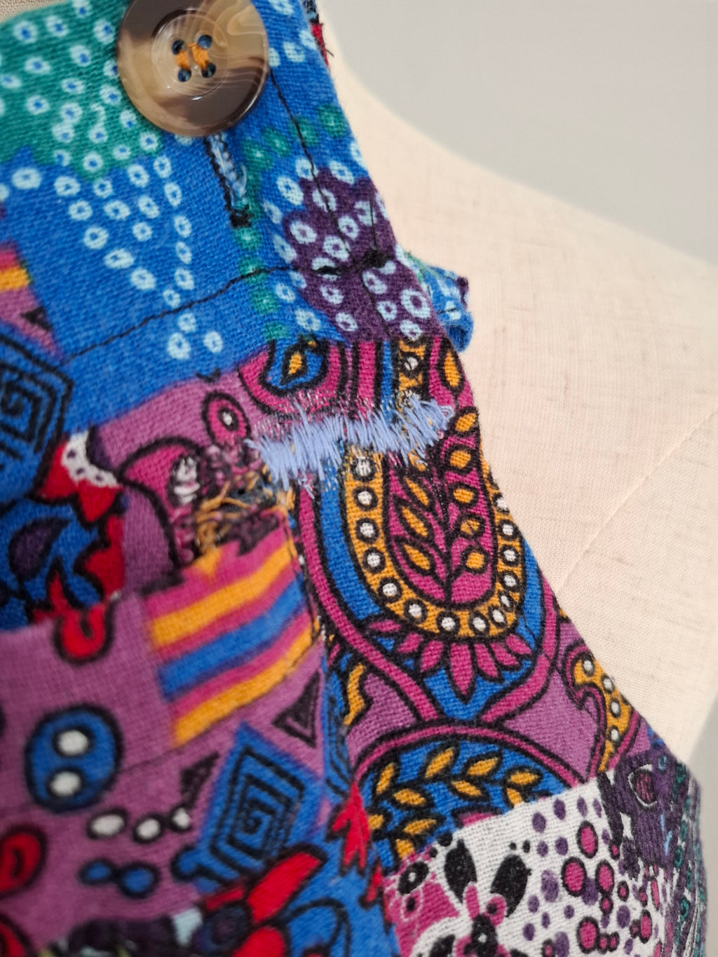 Close-up of a colorful patchwork fabric garment with a brown button, featuring vibrant blue, pink, purple, yellow, and red patterns on a light beige mannequin torso.