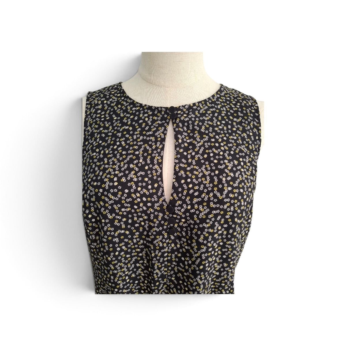 A sleeveless black top with a small beige and white floral pattern, displayed on a cream-colored mannequin torso against a white background.