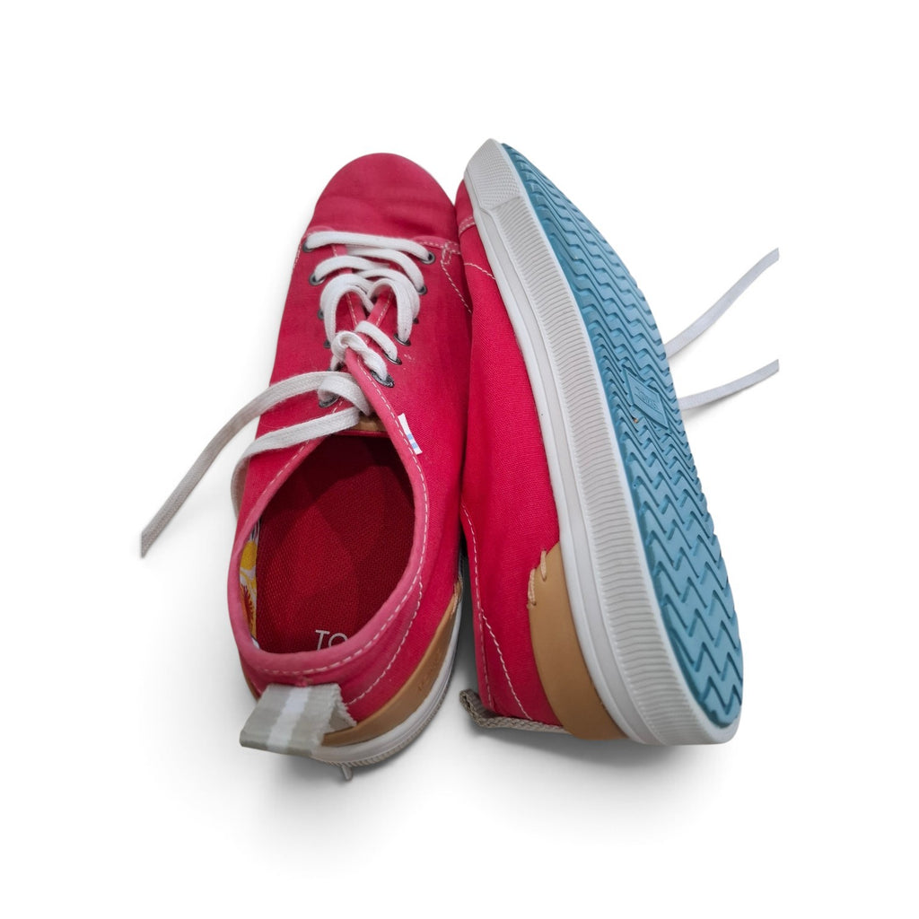 A pair of low-top canvas sneakers in a vibrant pinkish-red color, featuring white laces, a white rubber midsole, a tan suede heel tab, and a light blue textured outsole.