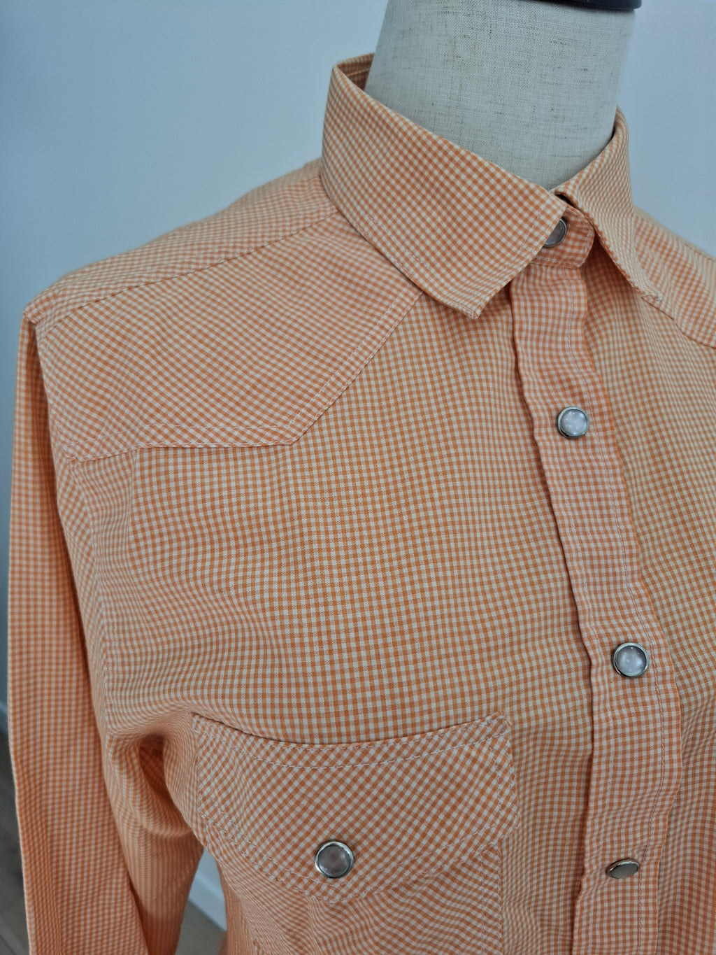 A close-up of a women's long-sleeve button-up shirt on a mannequin torso, featuring a fine orange and white gingham pattern, western-style chest pockets with snap closures, and silver snap buttons.