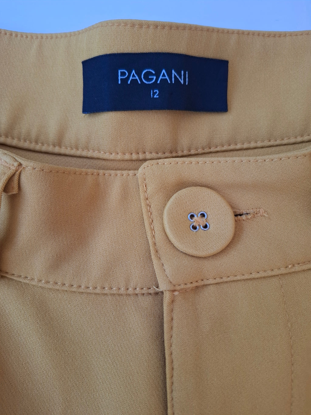 Close-up of tan trousers showing the waistband, a dark blue PAGANI size 12 label, and a matching tan button.