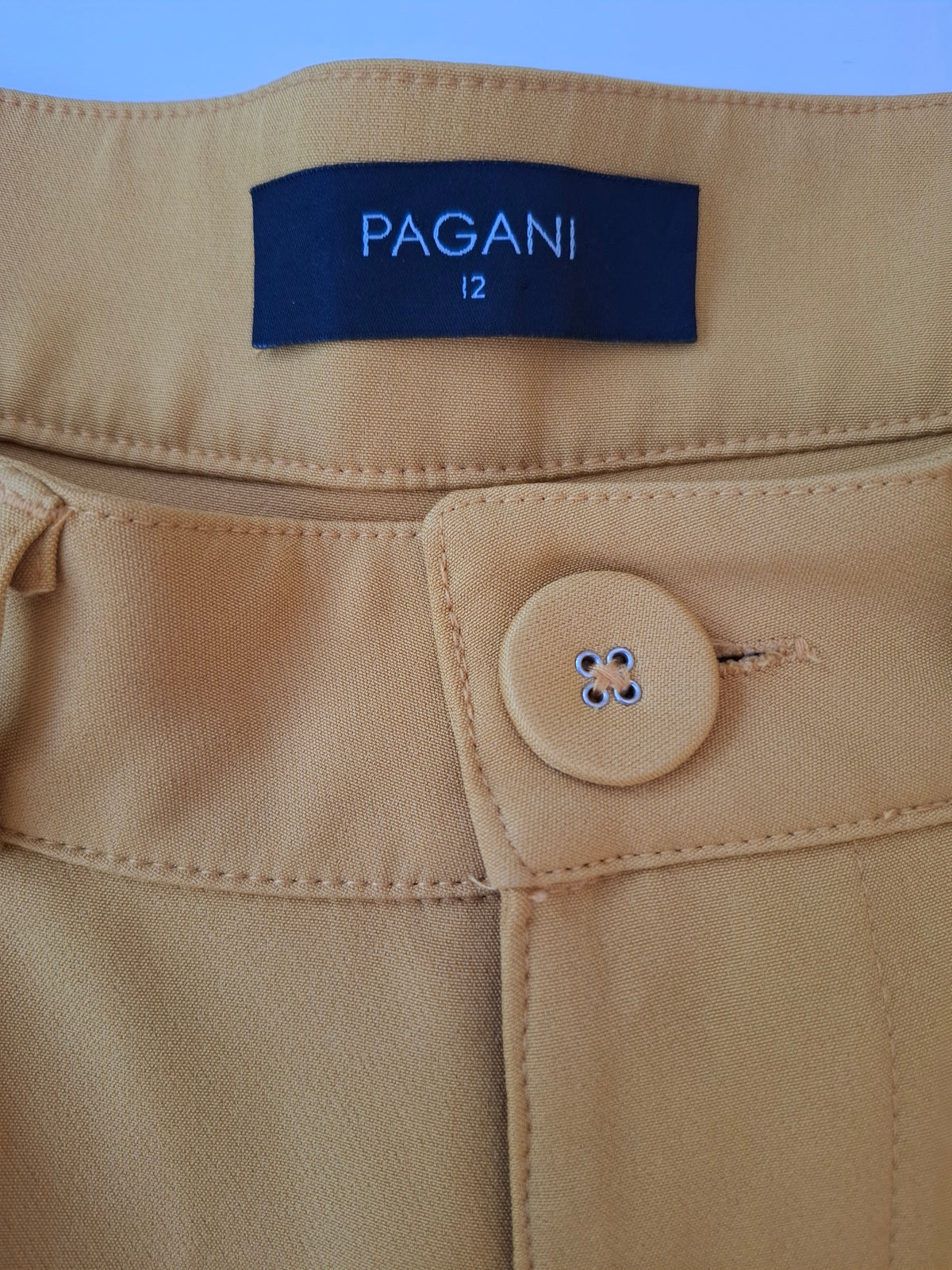 Close-up of tan trousers showing the waistband, a dark blue PAGANI size 12 label, and a matching tan button.