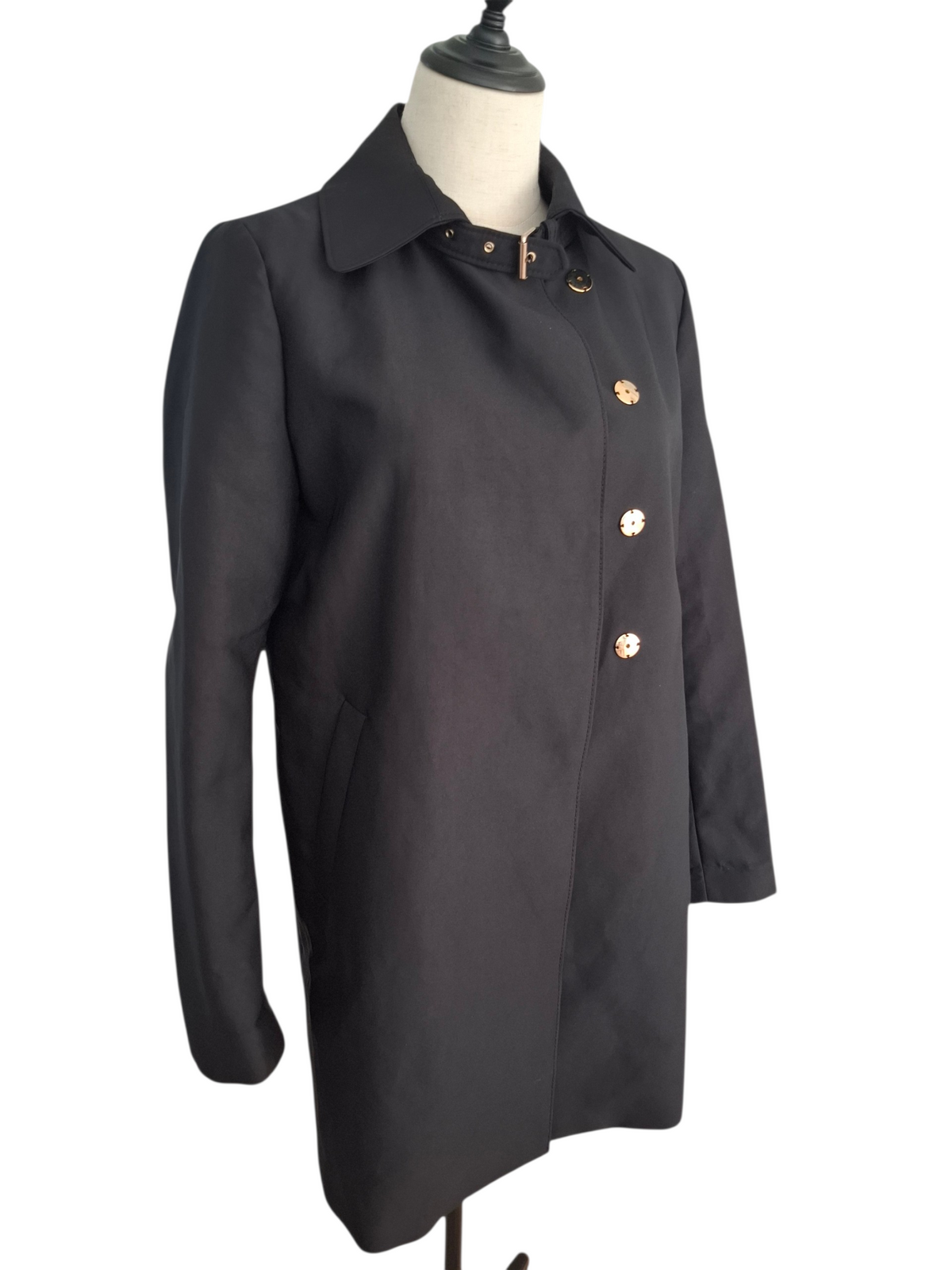 A black longline coat with gold-tone buttons, displayed on a cream mannequin torso against a white background.