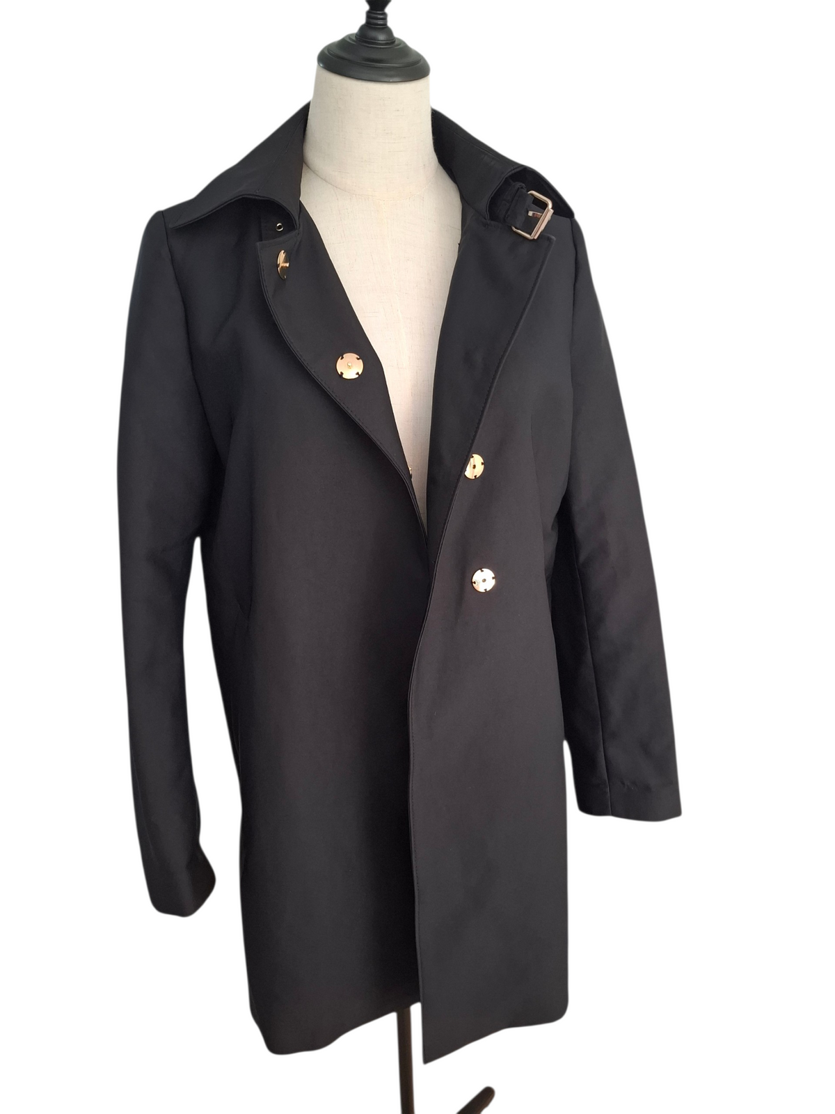 A black double-breasted trench coat with gold-tone buttons and a matching belt buckle, displayed on a cream mannequin torso against a white background.