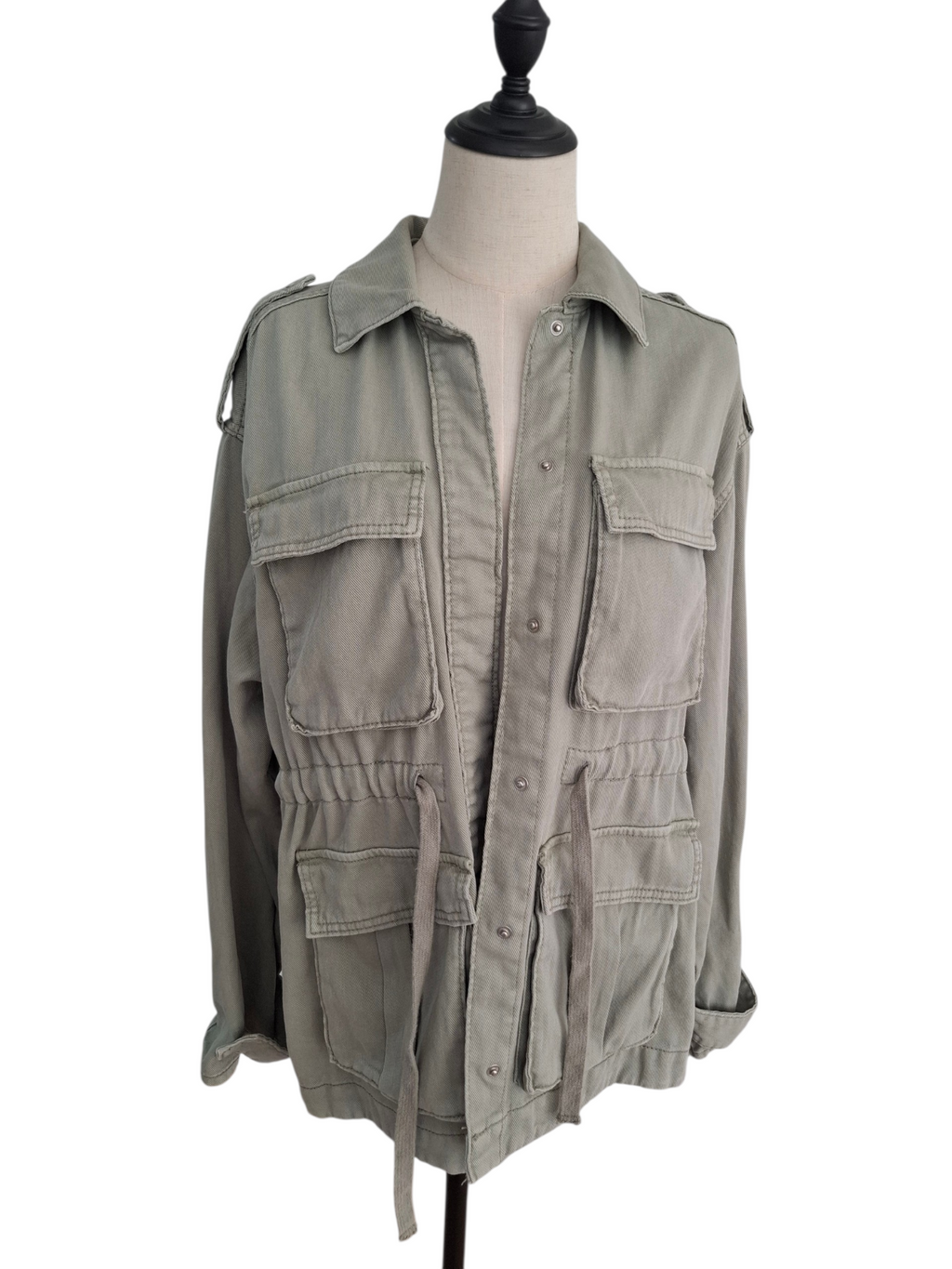 A light olive green military-style utility jacket displayed on a cream-colored dress form mannequin against a plain white background.