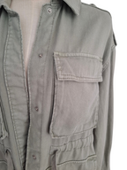 Close-up of a light gray utility shirt jacket with a button-down front, chest pocket, and drawstring waist, shown on a mannequin torso against a white background.