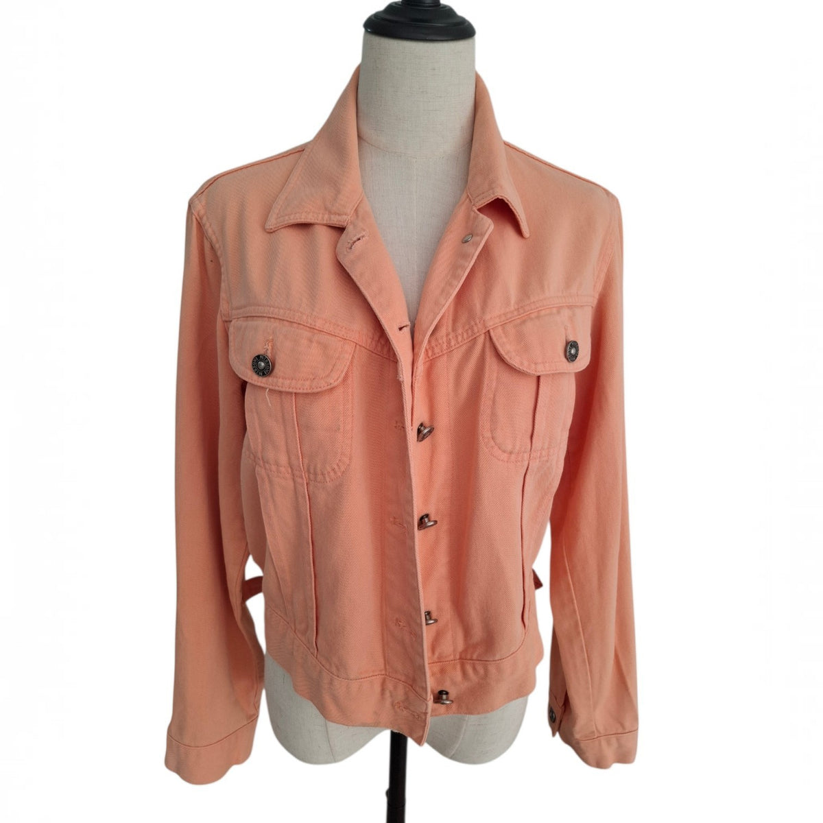 A peach-colored cropped denim jacket with a collar, button front, and two chest flap pockets, displayed on a dress form mannequin against a white background.