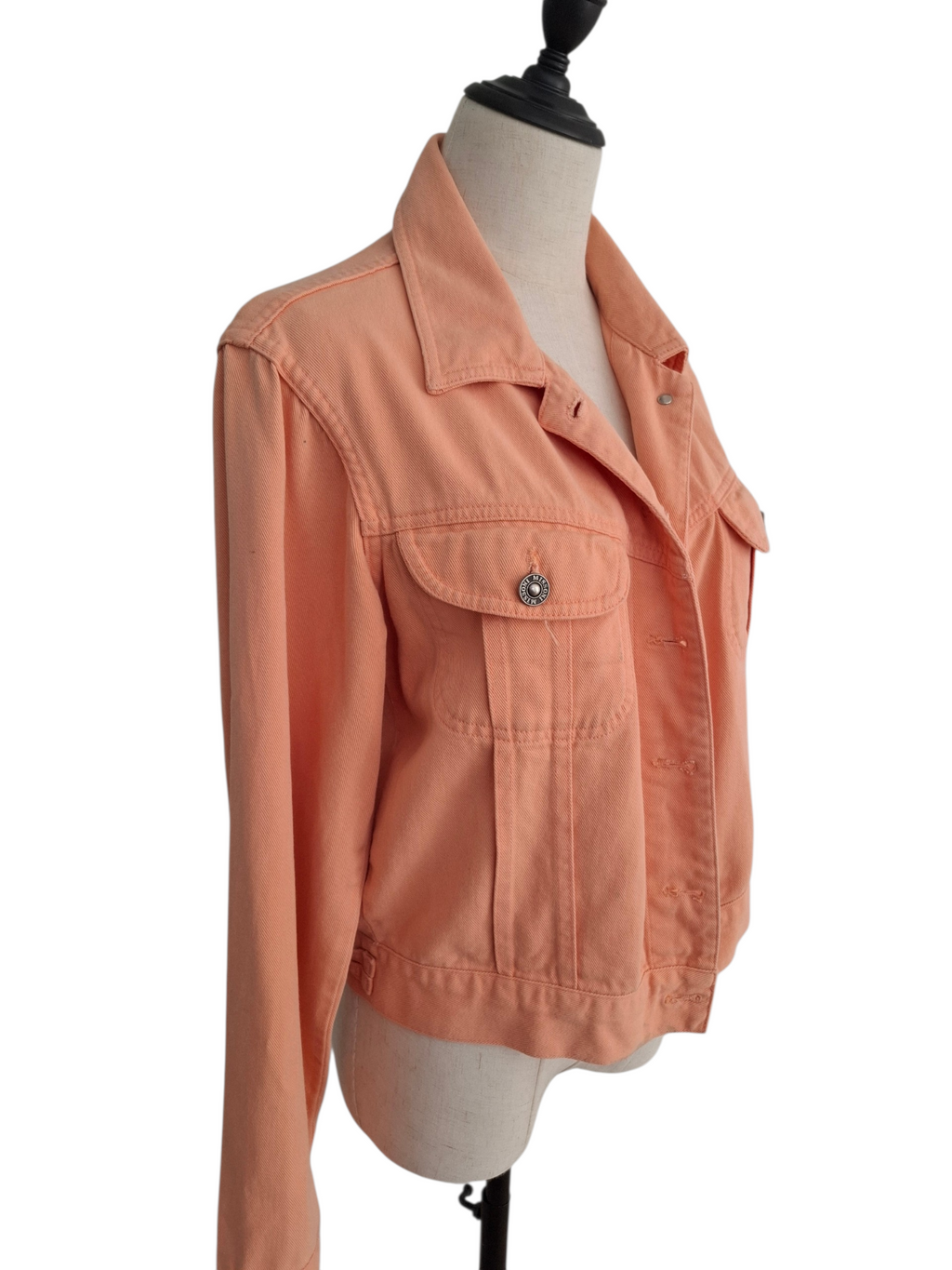 A cropped, long-sleeve denim jacket in a muted peach-orange color, displayed on a beige fabric mannequin torso against a plain white background.