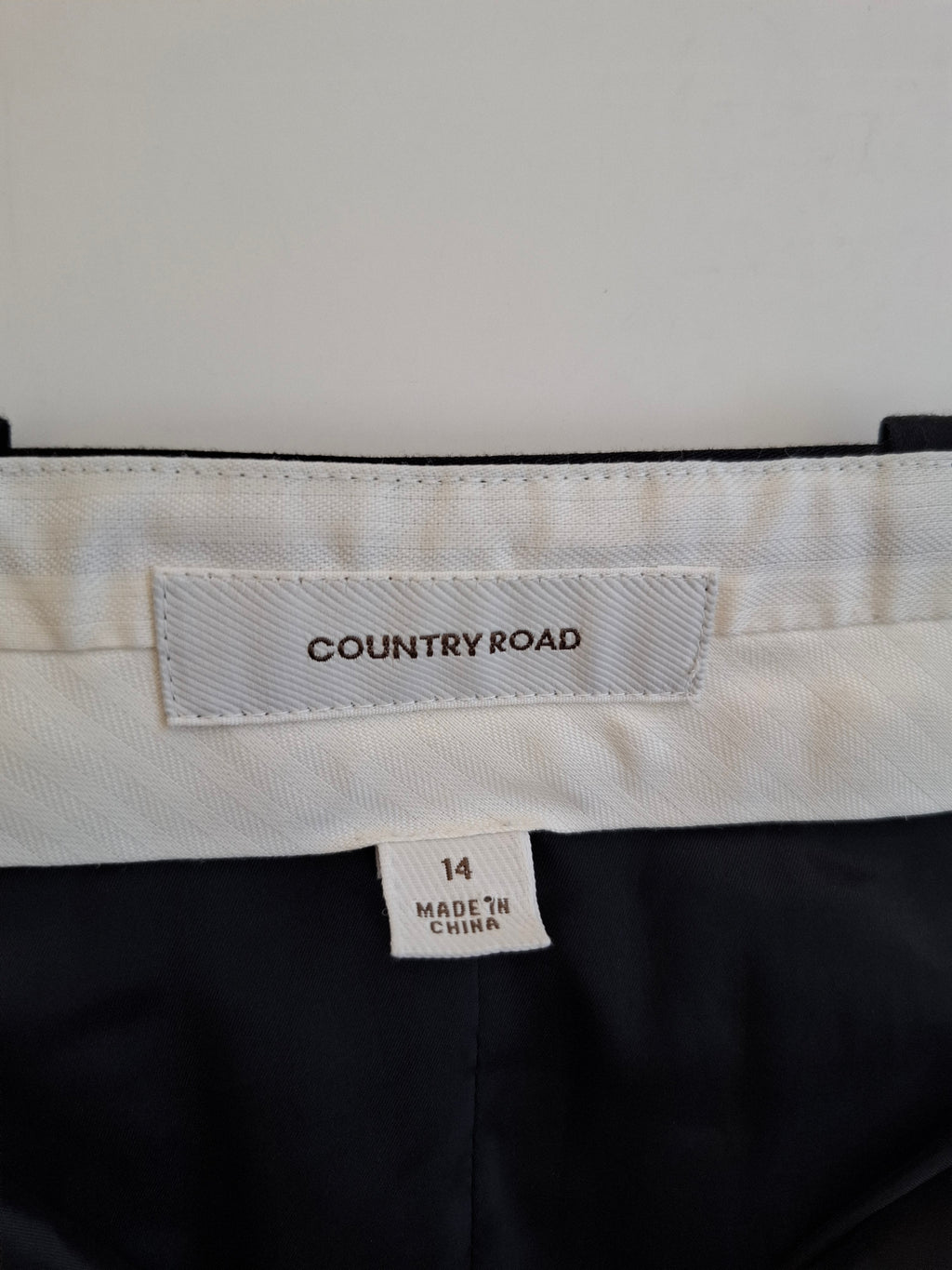 Close-up of the waistband of Country Road trousers, showing a white fabric label with the brand name 'COUNTRY ROAD' and a size tag indicating '14 MADE IN CHINA'. The trousers are black with a white waistband.