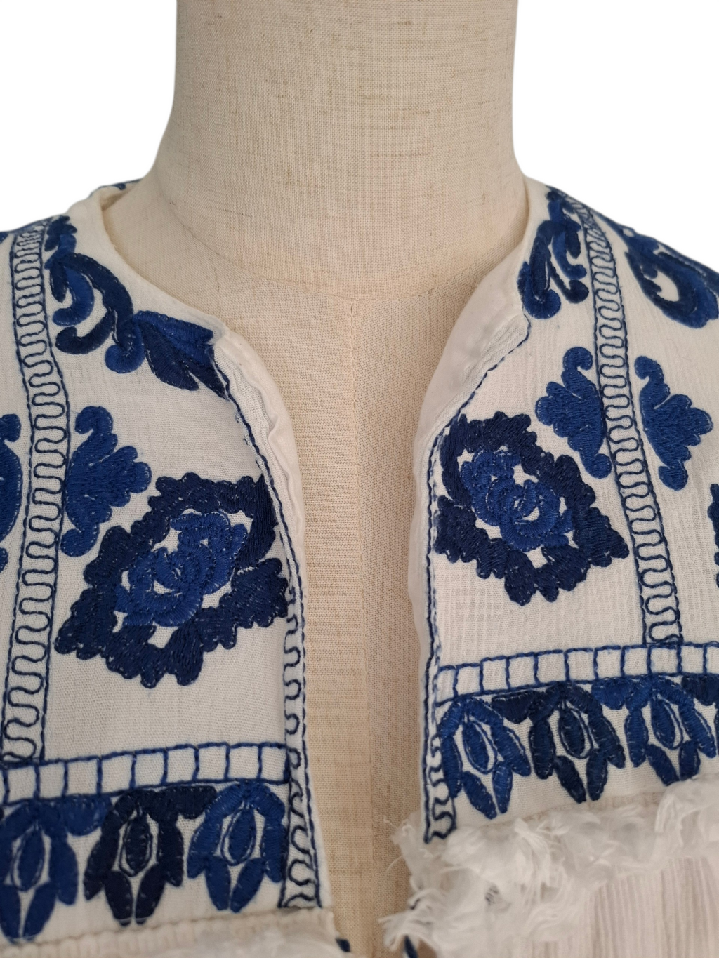 Close-up of a cream-colored embroidered garment displayed on a beige fabric mannequin torso, featuring dark blue floral embroidery and fringed trim.