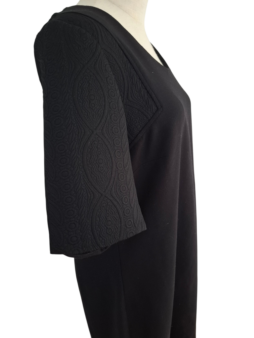 A black V-neck dress or tunic displayed on a mannequin, featuring textured, patterned elbow-length sleeves and a smooth black body, shown against a plain white background.