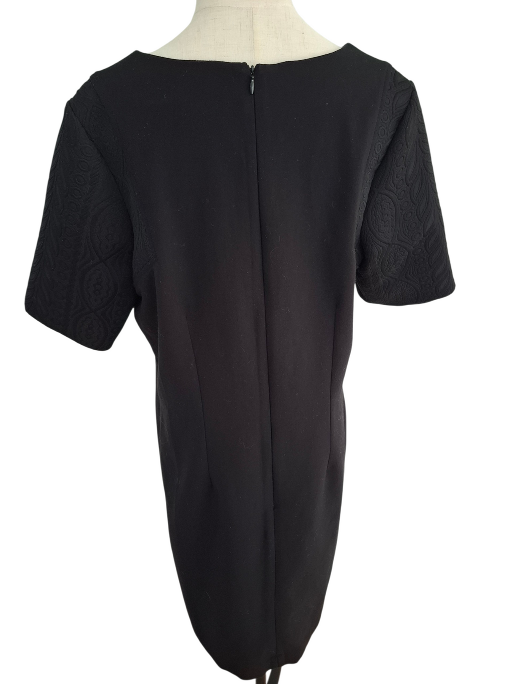 A black short-sleeve dress shown from the back on a mannequin, featuring a central zipper and textured patterned sleeves.
