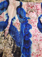 A close-up of a printed fabric featuring vivid blue peacocks over a pink floral background.