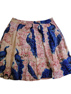 A pleated A-line skirt featuring a vibrant blue peacock and cherry blossom print on a light beige background.