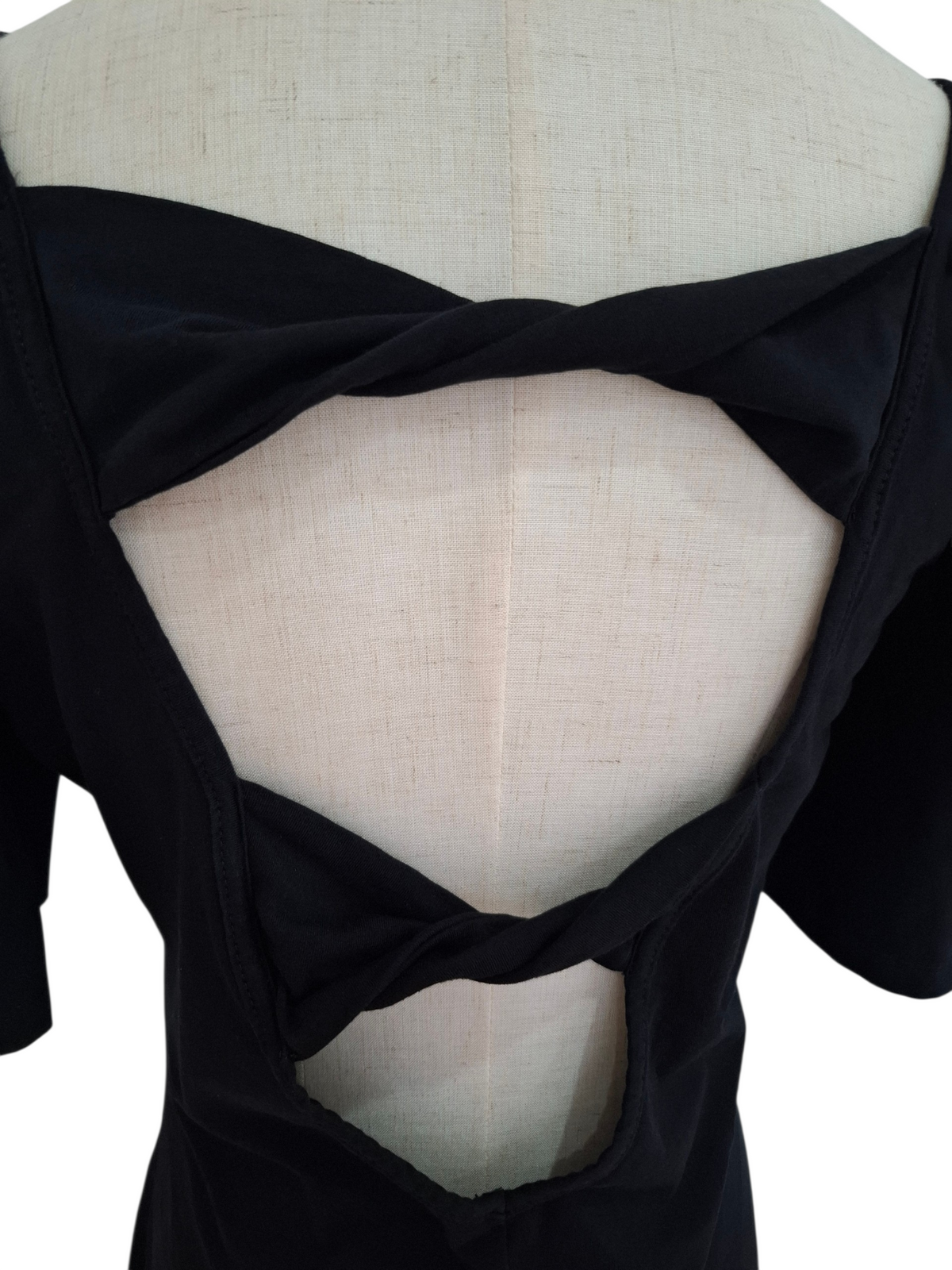 Close-up back view of a black short-sleeve top displayed on a cream fabric dress form, showing a large open back with twisted crossover straps.