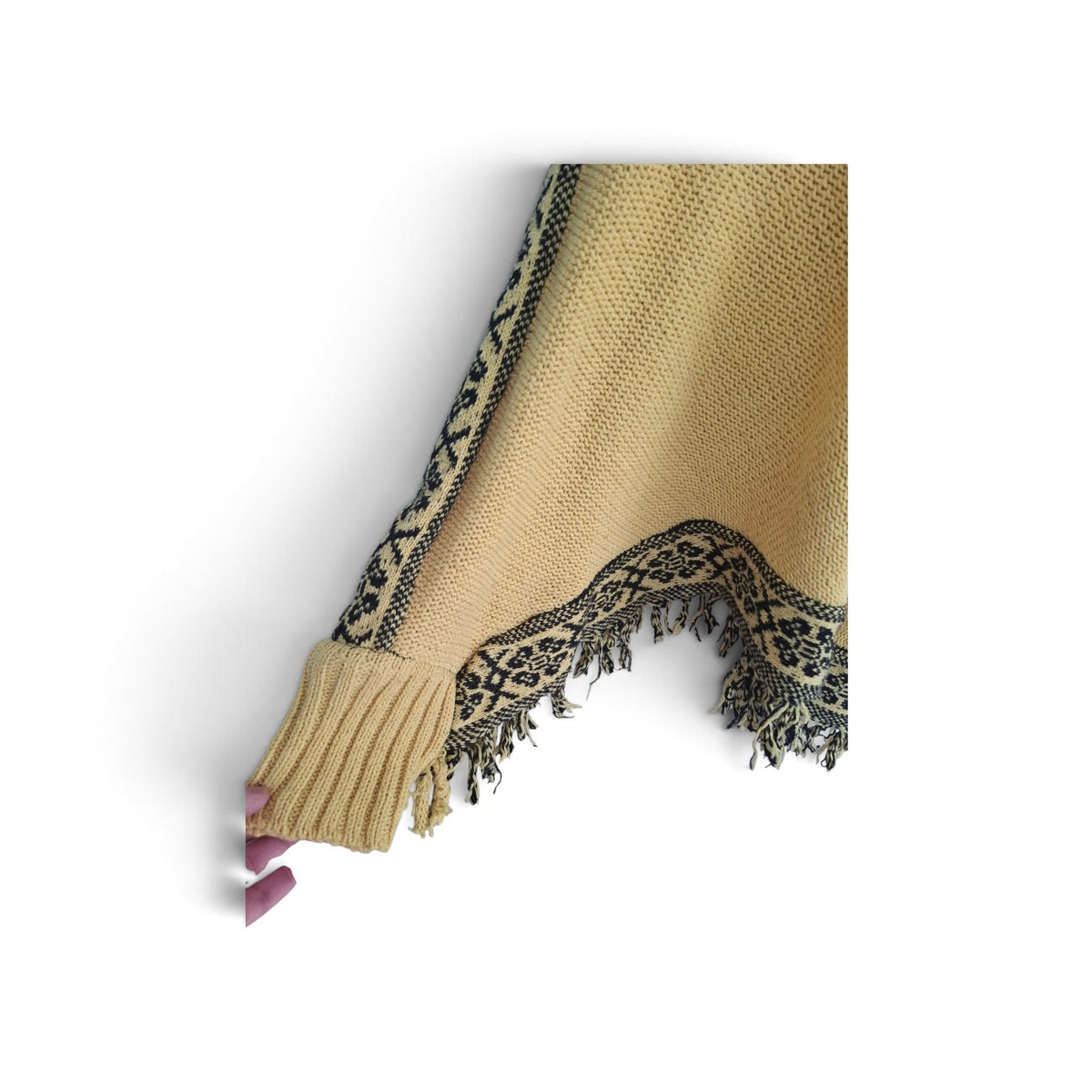 A close-up of a beige knit poncho sweater, showing a ribbed cuff and a black patterned fringe trim on a white background.