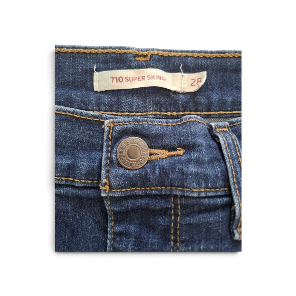 Close-up of dark blue denim jeans showing the waistband, a cream-colored '710 SUPER SKINNY' size 28 label, and a bronze-toned metal button on the front pocket.