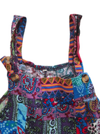 A close-up of a colorful patchwork-print garment, likely a dress or romper, featuring a busy pattern of paisley, stripes, and geometric designs in purple, blue, red, orange, and green. It has a solid white background.