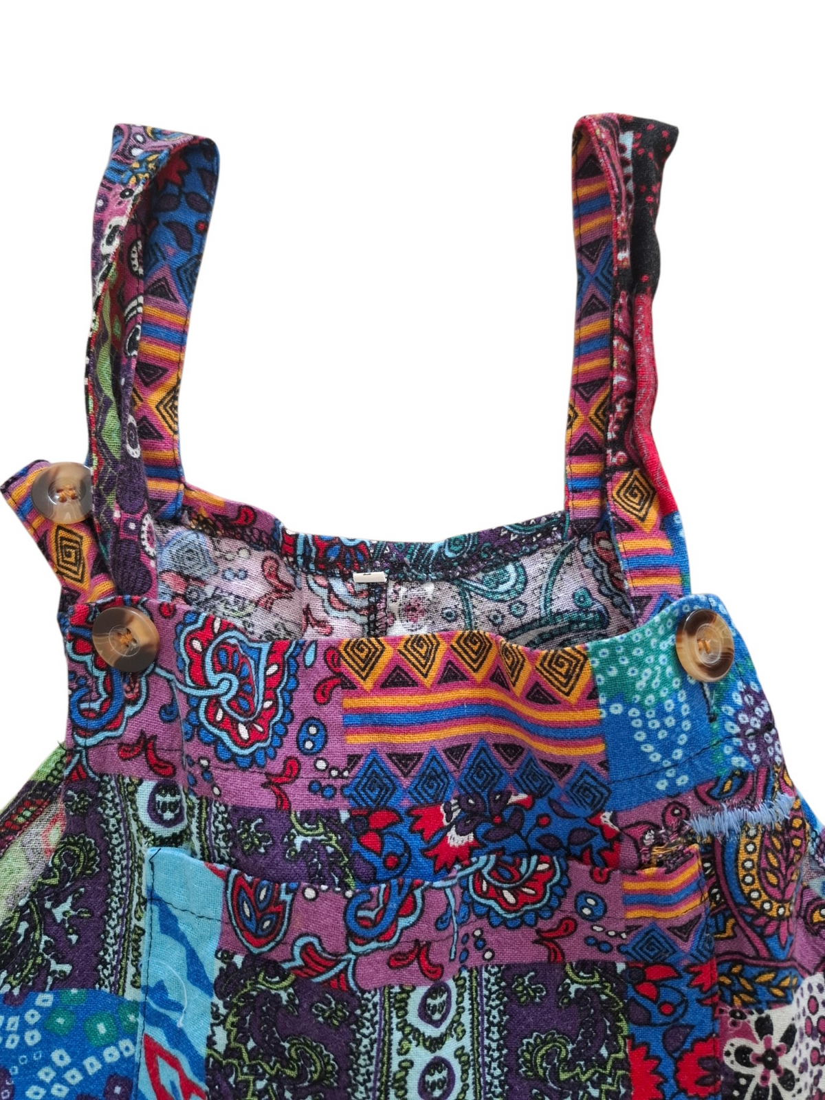 A close-up of a colorful patchwork-print garment, likely a dress or romper, featuring a busy pattern of paisley, stripes, and geometric designs in purple, blue, red, orange, and green. It has a solid white background.