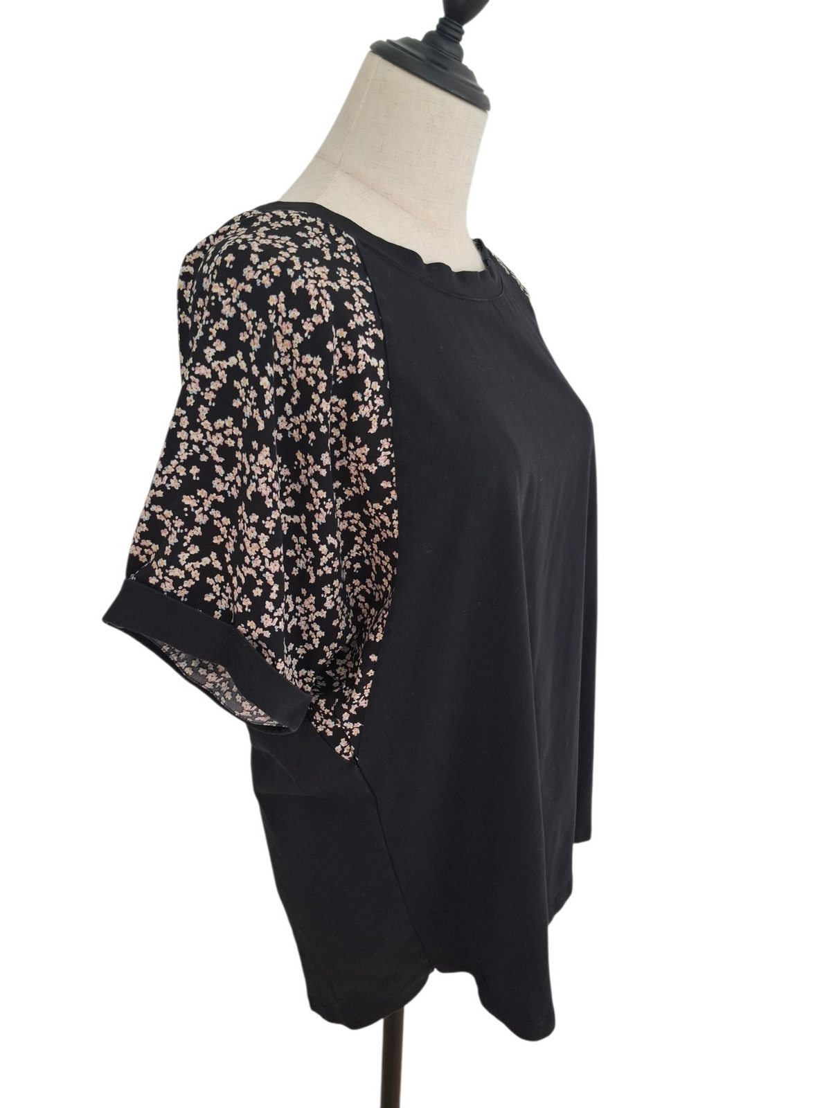 A black short-sleeve top with sheer floral-patterned sleeves, displayed on a cream dress form against a white background.