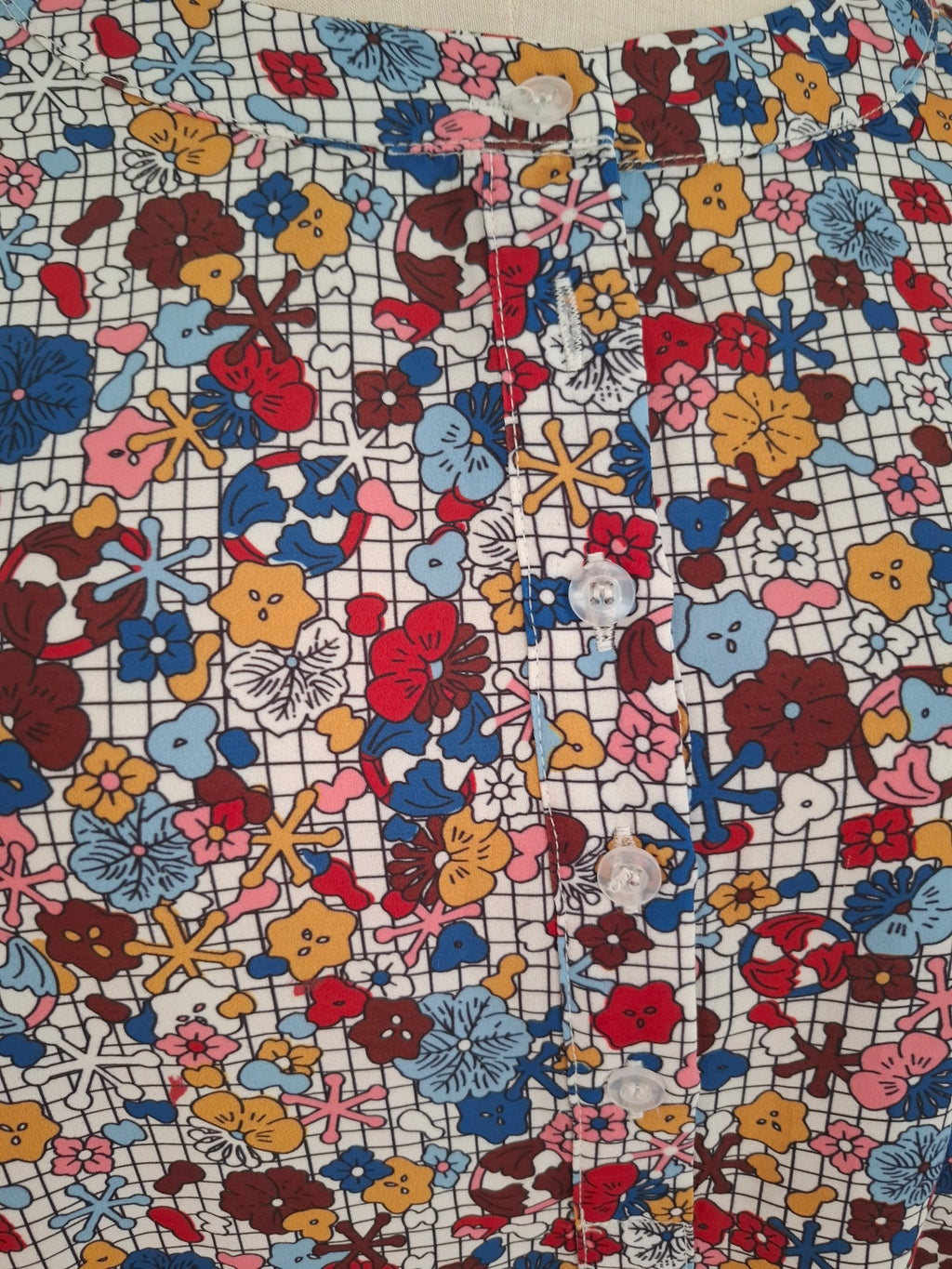 A close-up of a colorful floral fabric with a white grid background and multicolored flowers, featuring white buttons sewn along the front.