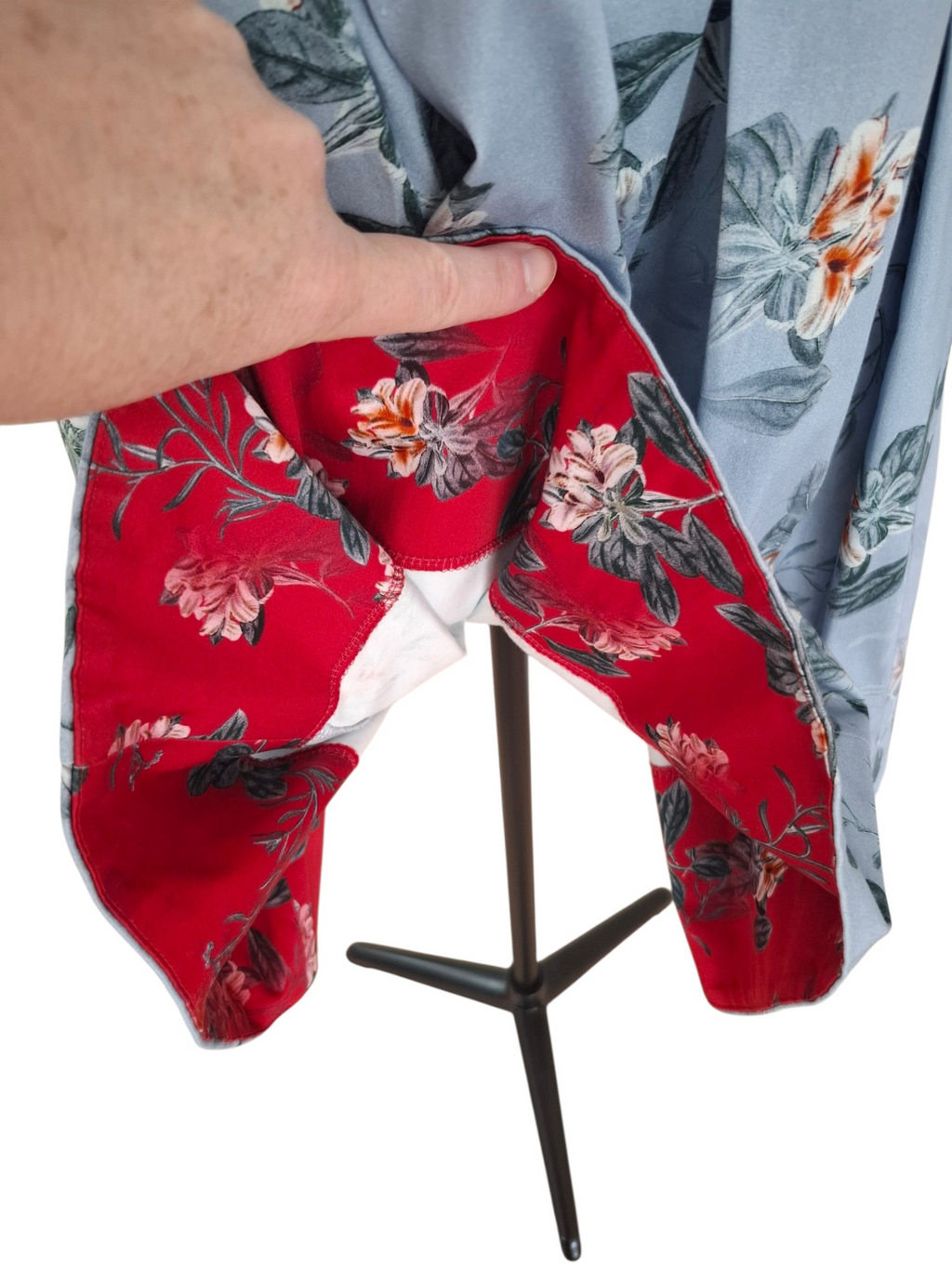 A close-up of a garment displayed on a black stand, showing a light blue outer fabric with a floral print and a bright red inner lining with a larger floral pattern. A human hand is pulling open the garment to reveal the lining, against a plain white background.