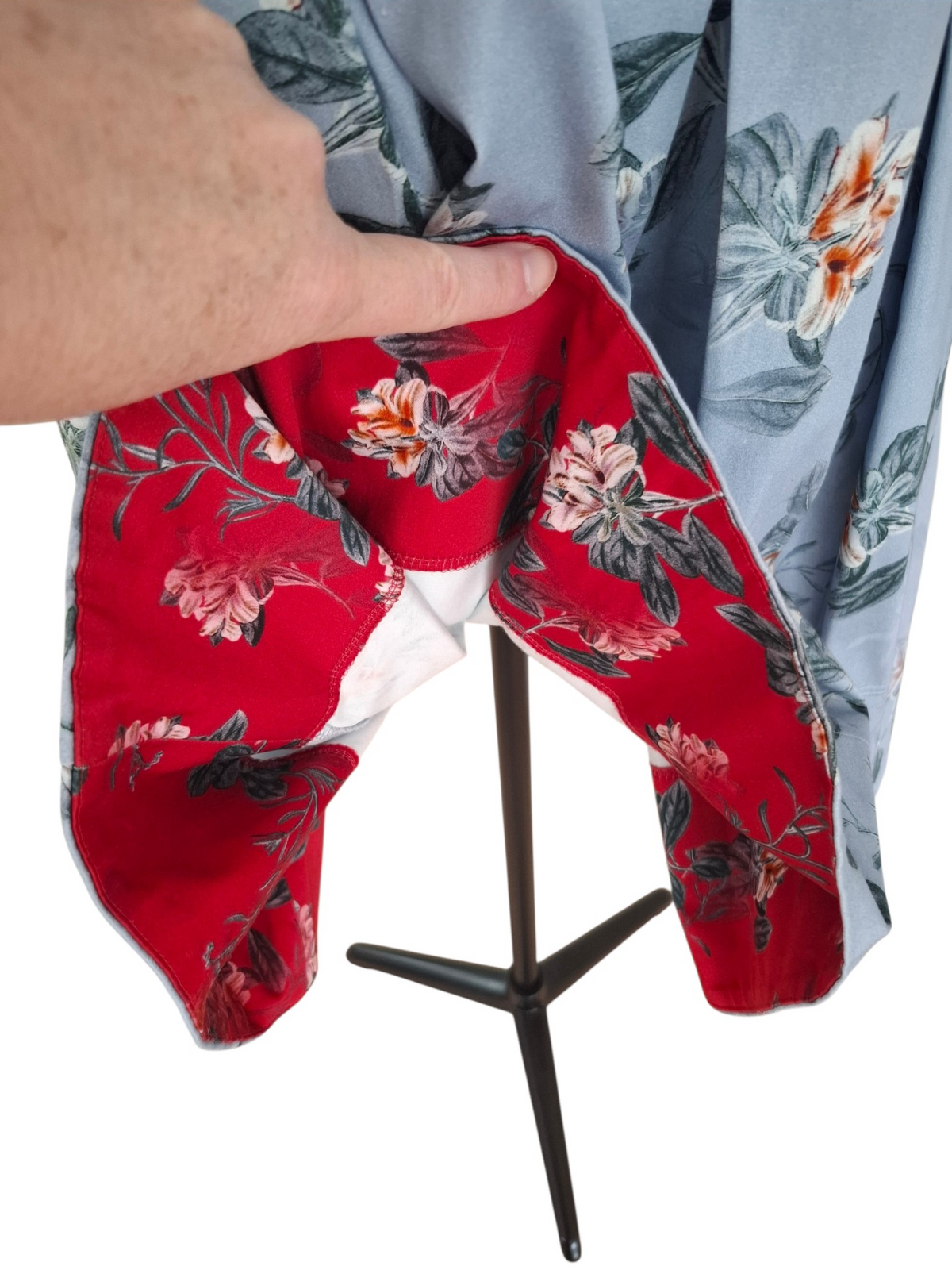 A close-up of a garment displayed on a black stand, showing a light blue outer fabric with a floral print and a bright red inner lining with a larger floral pattern. A human hand is pulling open the garment to reveal the lining, against a plain white background.