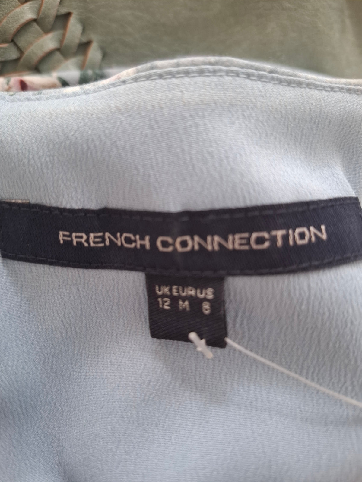 Close-up of a garment's inner neck label reading 'FRENCH CONNECTION' in white text on a black band, with a size tag below and a light blue fabric background.