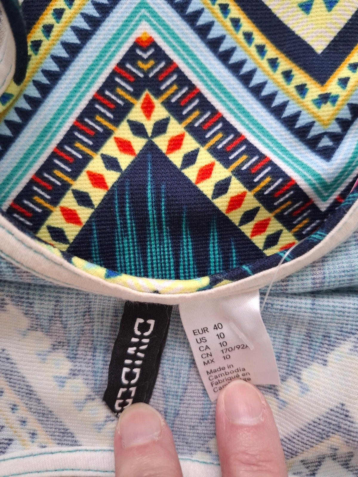 Close-up of a patterned garment interior with a visible sewn-in brand tag reading DIVIDES and a white size tag, with two fingers pointing at the size information.