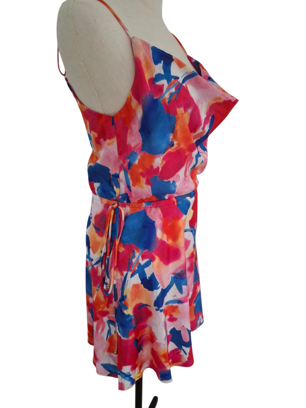 A sleeveless wrap-style dress with a vibrant abstract watercolor print in pink, blue, orange, and white, displayed on a beige dress form mannequin against a white background.
