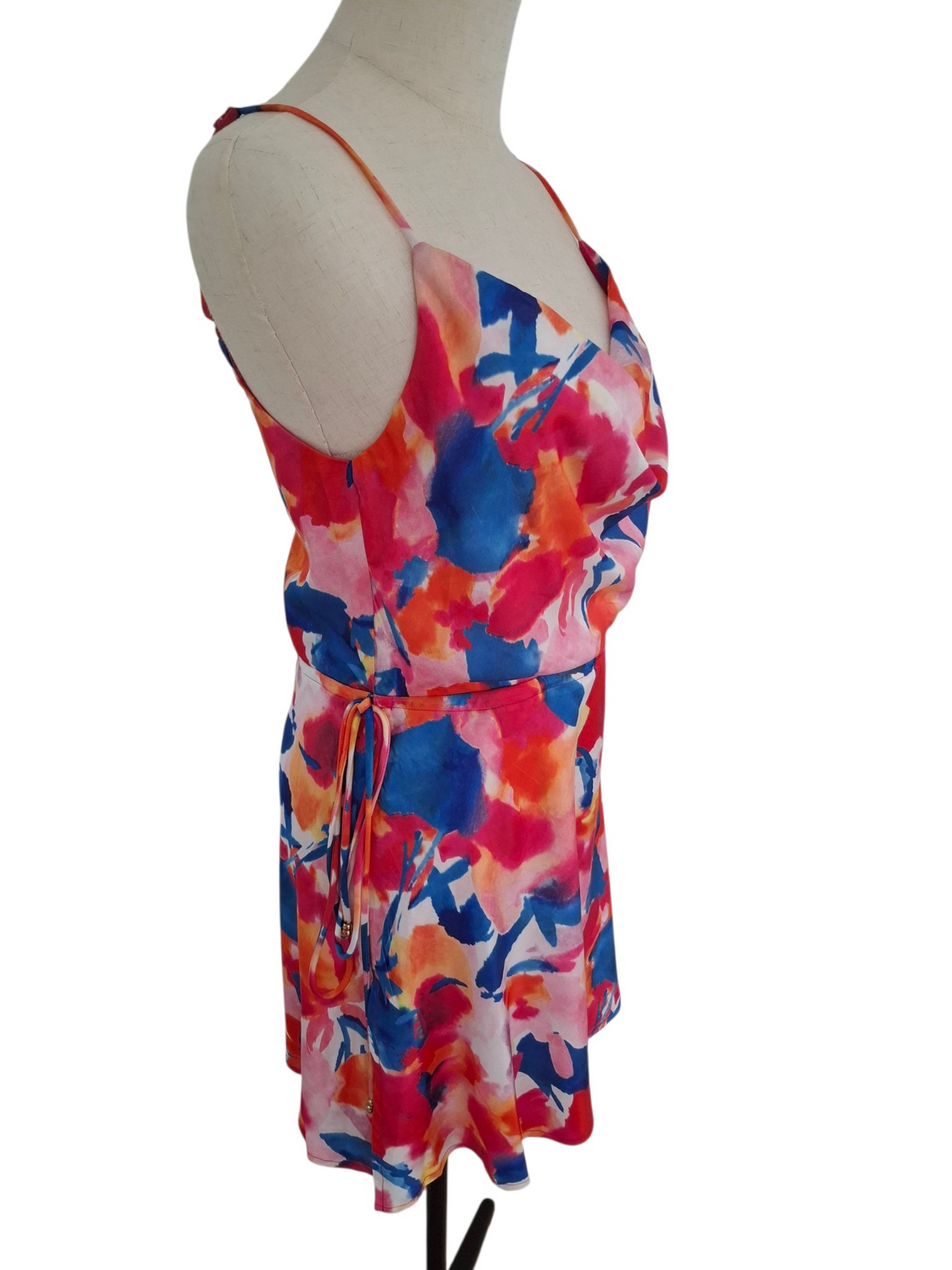 A sleeveless wrap-style dress with thin straps and a tie waist, featuring a vibrant abstract watercolor floral print in pink, blue, orange, and white, displayed on a mannequin torso against a white background.