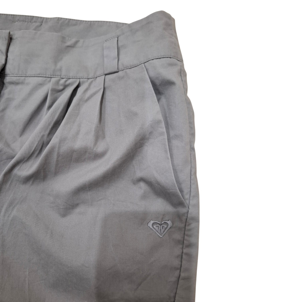 Close-up of gray pleated shorts with a small white embroidered Quiksilver logo on the lower leg, set against a plain white background.