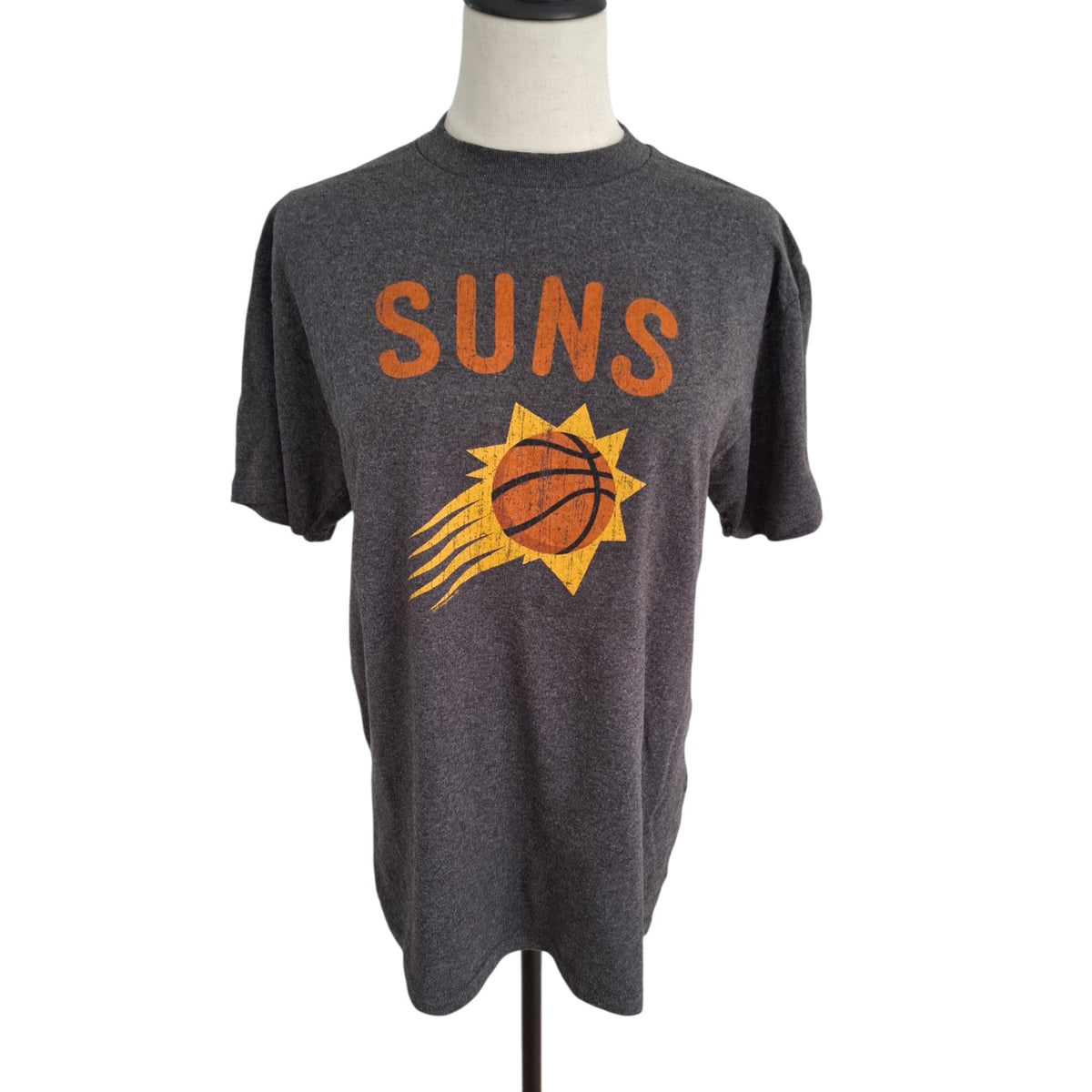 A dark heather gray short-sleeve t-shirt displayed on a mannequin torso, featuring a distressed orange 'SUNS' text and a yellow and orange Phoenix Suns basketball logo on the front.