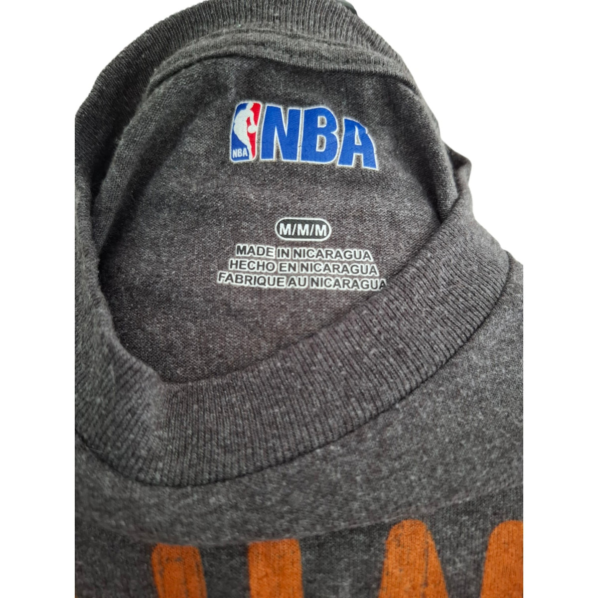 Close-up of the inside neck of a dark gray heathered sweatshirt featuring a blue and red embroidered NBA logo on the inner collar, with size text 'M/M/N' and 'MADE IN NICARAGUA' printed below it.