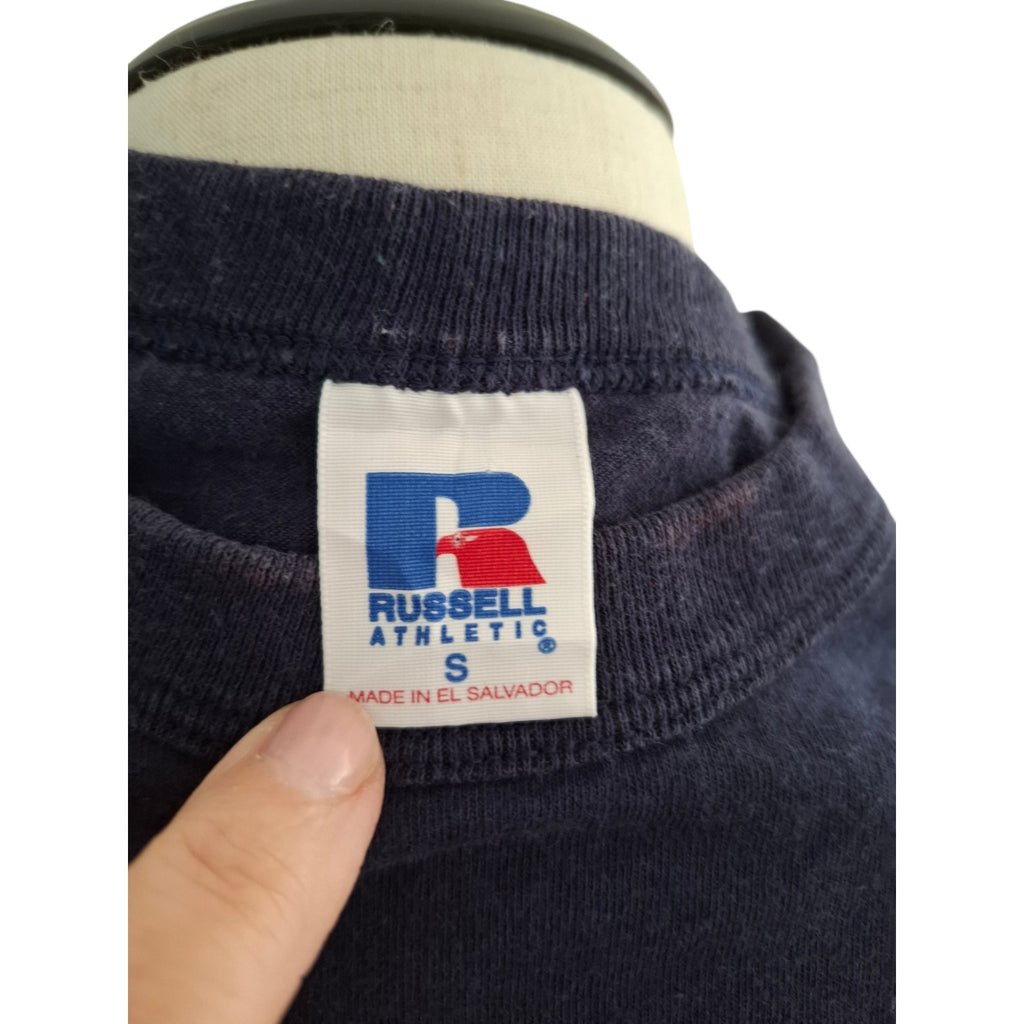 Close-up of a navy blue Russell Athletic sweatshirt on a mannequin, showing the neck label and a finger pointing at it.