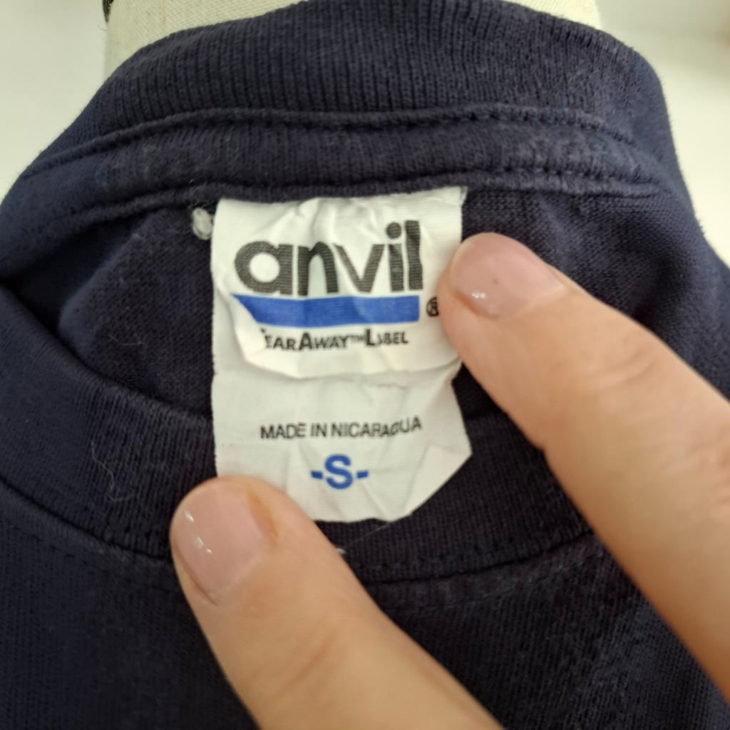 Close-up of a navy blue garment neck label with a finger pointing to the white Anvil brand tag, which reads 'TAR-AWAY-LISEL' and 'MADE IN NICARAGUA'.