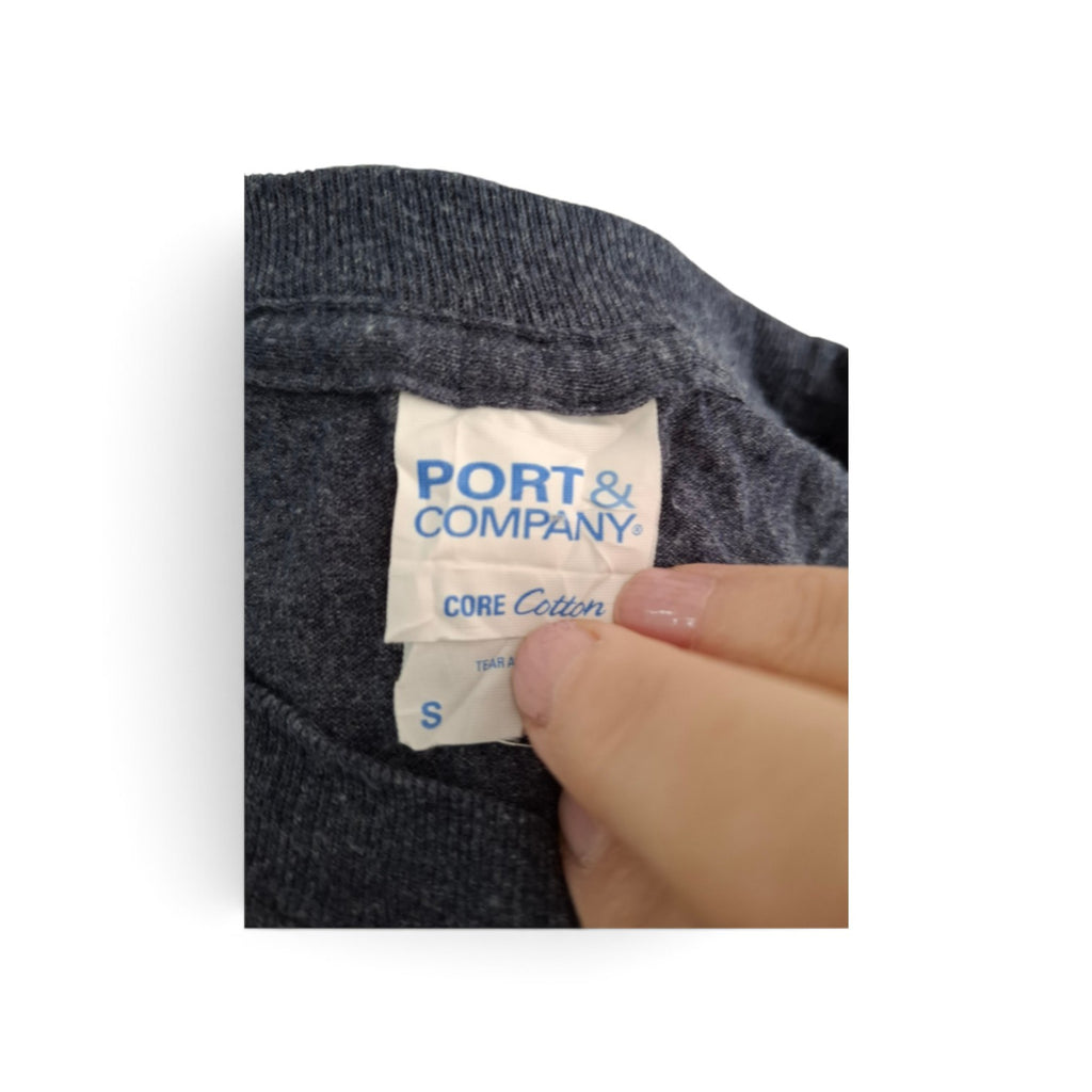 Close-up of a dark gray heather knit garment neck label reading PORT & COMPANY and COTTON, with a hand holding the tag in place against a white background.