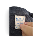 Close-up of a dark gray heather knit garment neck label reading PORT & COMPANY and COTTON, with a hand holding the tag in place against a white background.