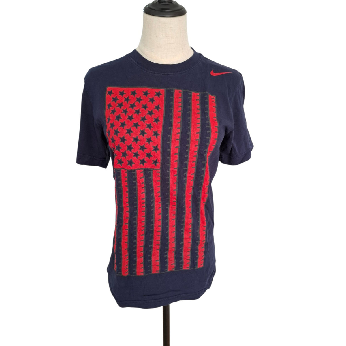 A navy blue Nike t-shirt displayed on a headless mannequin torso. The shirt features a large distressed red American flag graphic on the front, with a red Nike swoosh on the left chest.