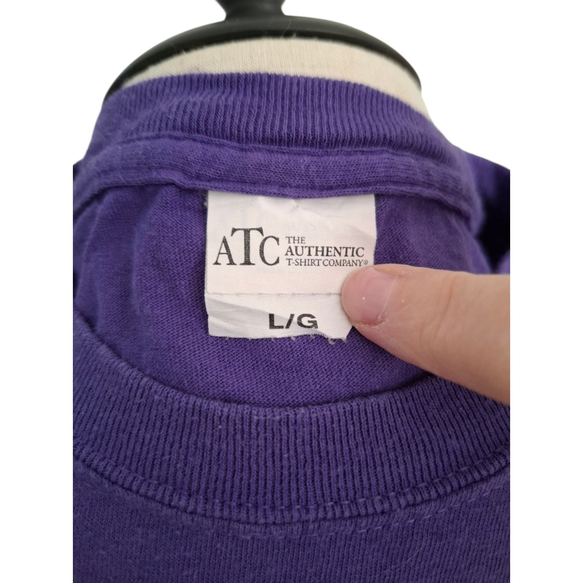 Close-up of a purple ribbed sweatshirt on a mannequin, showing the neck label for 'ATC The Authentic T-Shirt Company' and a finger pointing to the 'L/G' size marking.