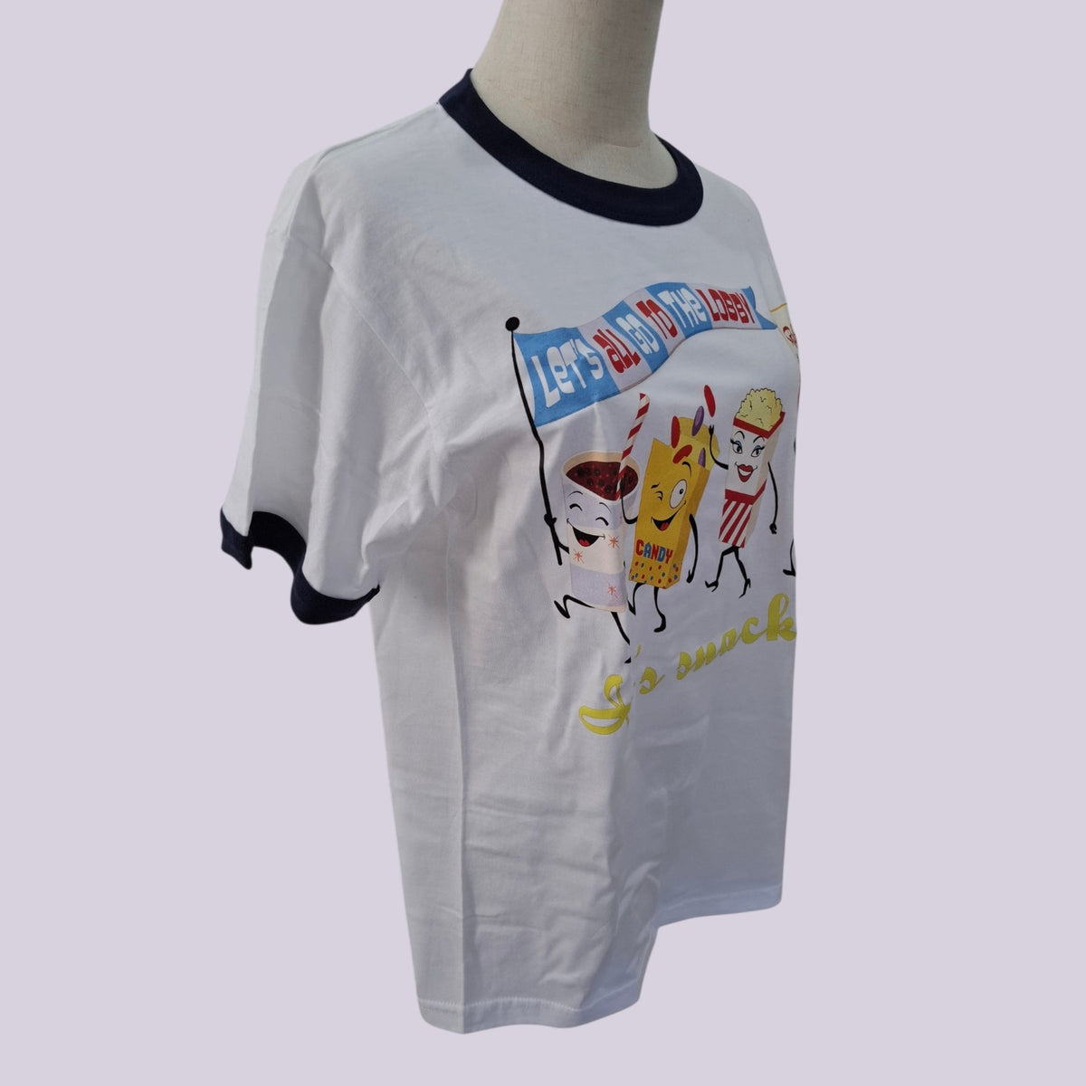 A white ringer t-shirt with black trim on the collar and sleeve cuffs, featuring a colorful cartoon graphic of snack characters on the front, displayed on a mannequin torso against a light gray background.