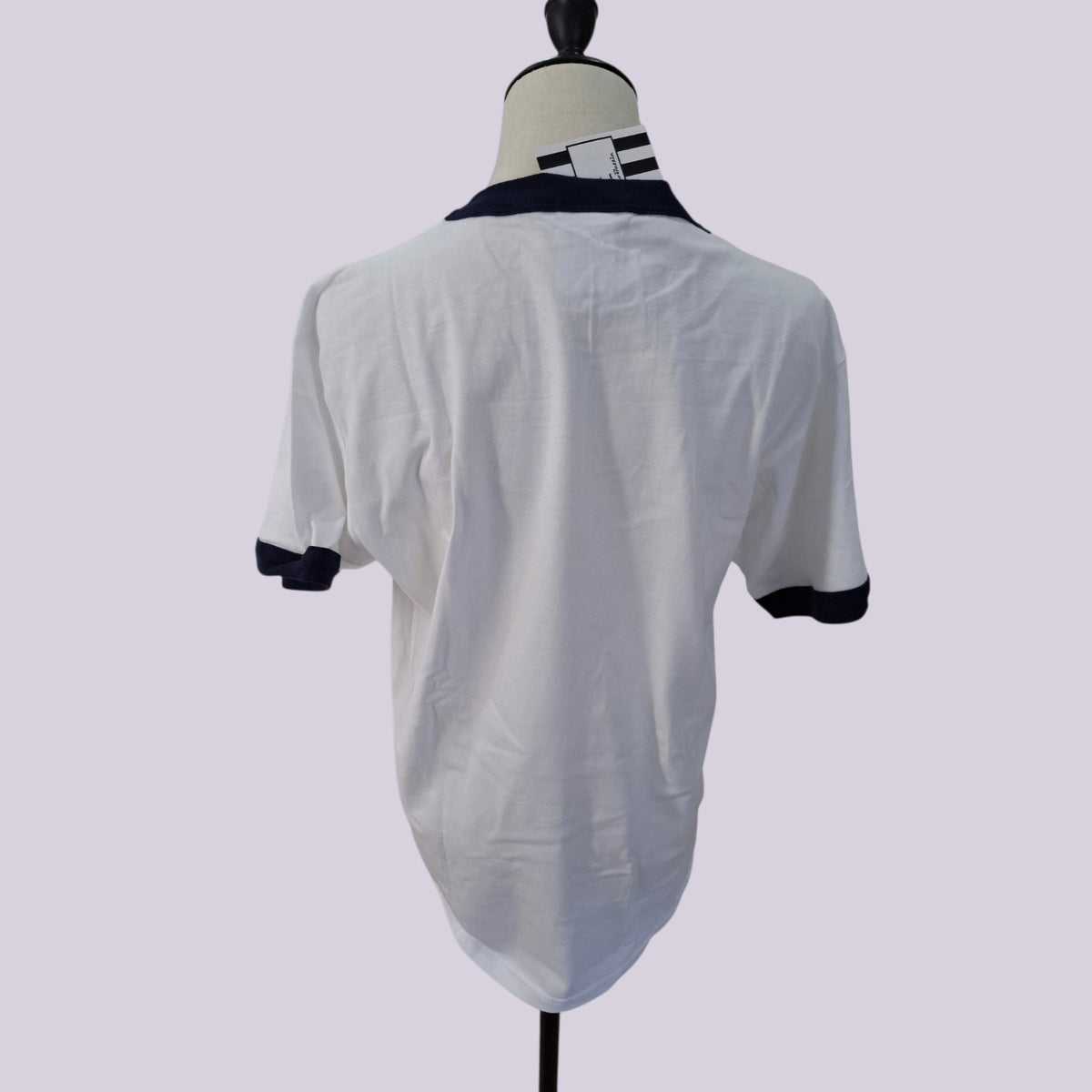 White short-sleeve ringer t-shirt with a dark navy collar and sleeve cuffs, displayed on a mannequin from the back against a light lavender background.