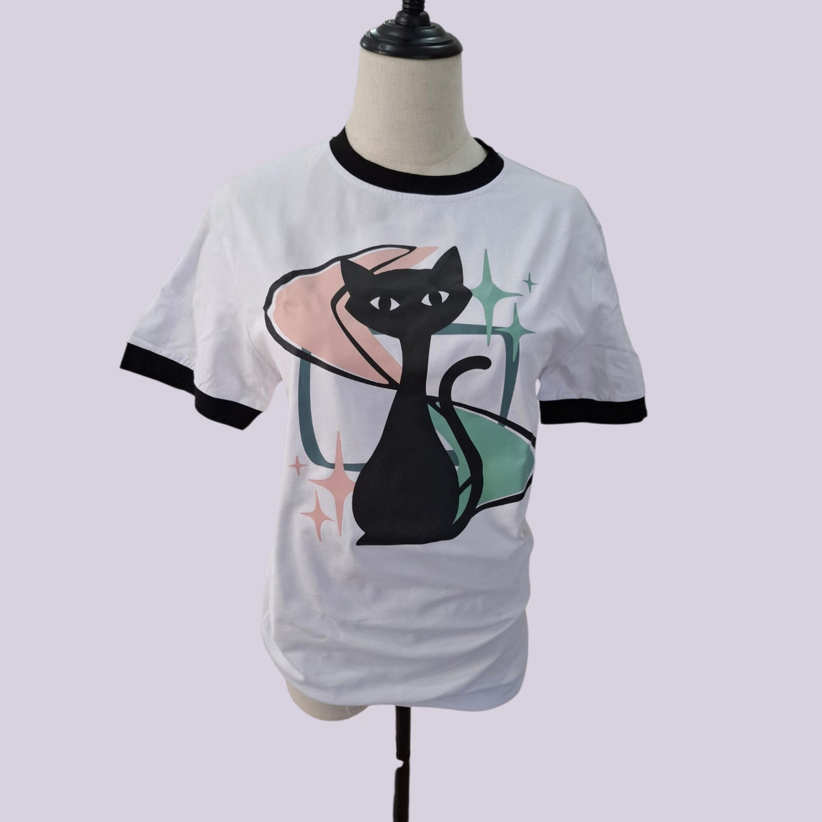 White ringer t-shirt with black trim on the collar and sleeves, featuring a graphic of a black cat sitting on a pink cushion with green sparkles, displayed on a mannequin torso.