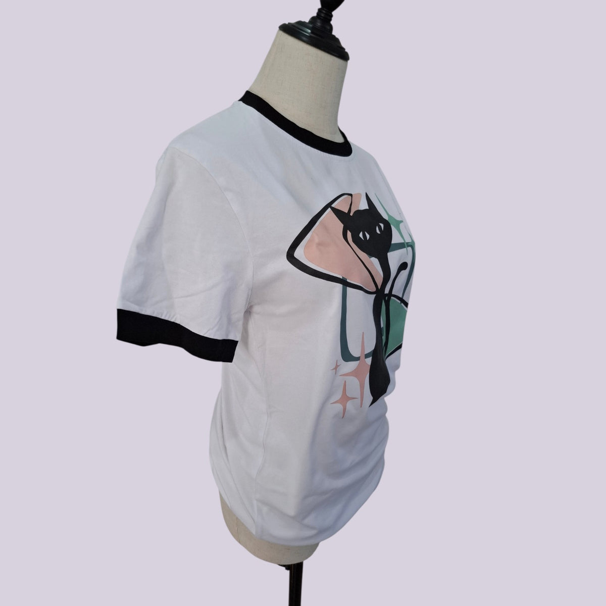 White ringer t-shirt with black trim on the collar and sleeves, featuring a graphic print of a black cat inside a pink and green tent, displayed on a mannequin torso against a light lavender background.
