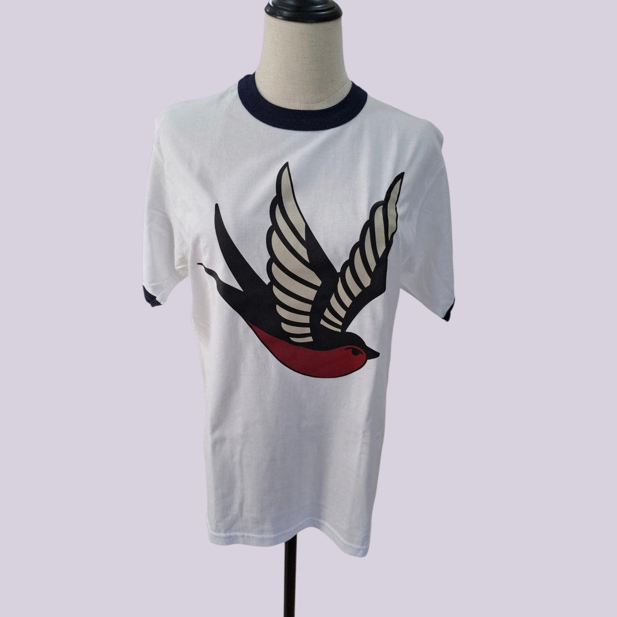 White ringer t-shirt on a mannequin torso, featuring a large stylized bird graphic on the front with black, cream, and red details, and a dark navy collar.