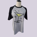 A gray and black raglan t-shirt displayed on a mannequin torso, featuring a graphic of a martini glass with an olive on a starry background.
