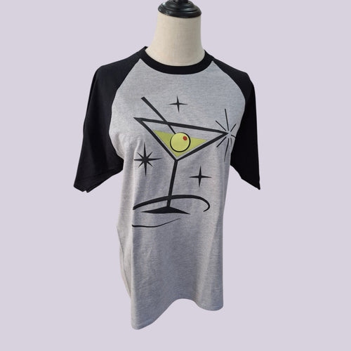A gray and black raglan t-shirt displayed on a mannequin torso, featuring a graphic of a martini glass with an olive on a starry background.