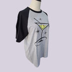 A heather gray and black raglan t-shirt displayed on a mannequin torso. The shirt features a graphic of a martini glass with a green olivine and black sparkles on the front, set against a solid light purple background.